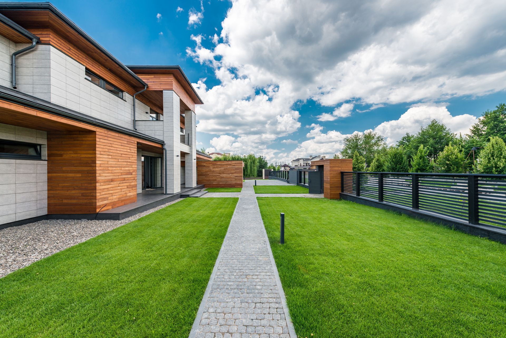 Scenery green yard of modern residential house with trees outside fence under blue cloudy sky